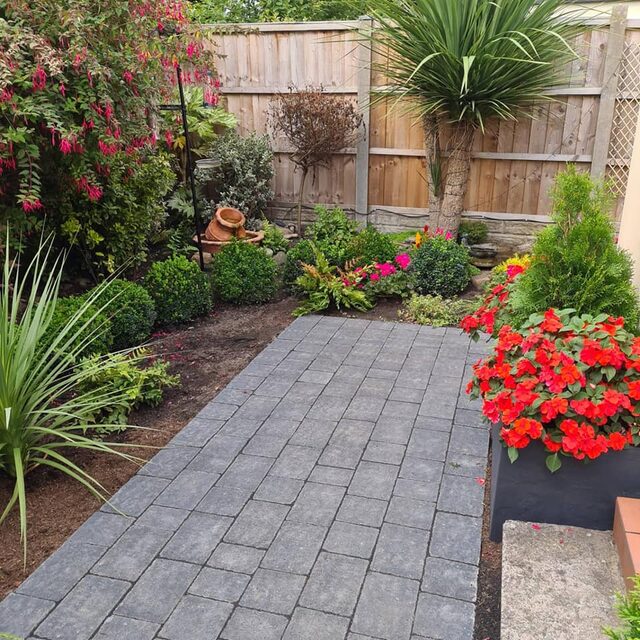 A block-paved garden path running between planted borders and a fuchsia hedge.
